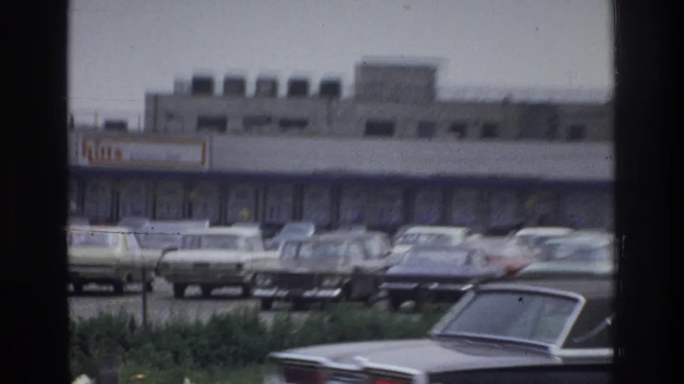 Soft 8mm frame from the Bronx showing rows of parked 1970s cars outside a shopping strip, partly framed by dark borders.