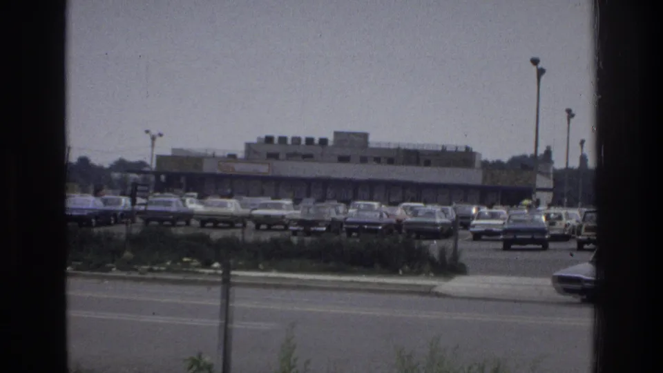 Wide 1975 Bronx home-movie view of a commercial parking lot, road lanes, streetlights, and rows of parked cars.