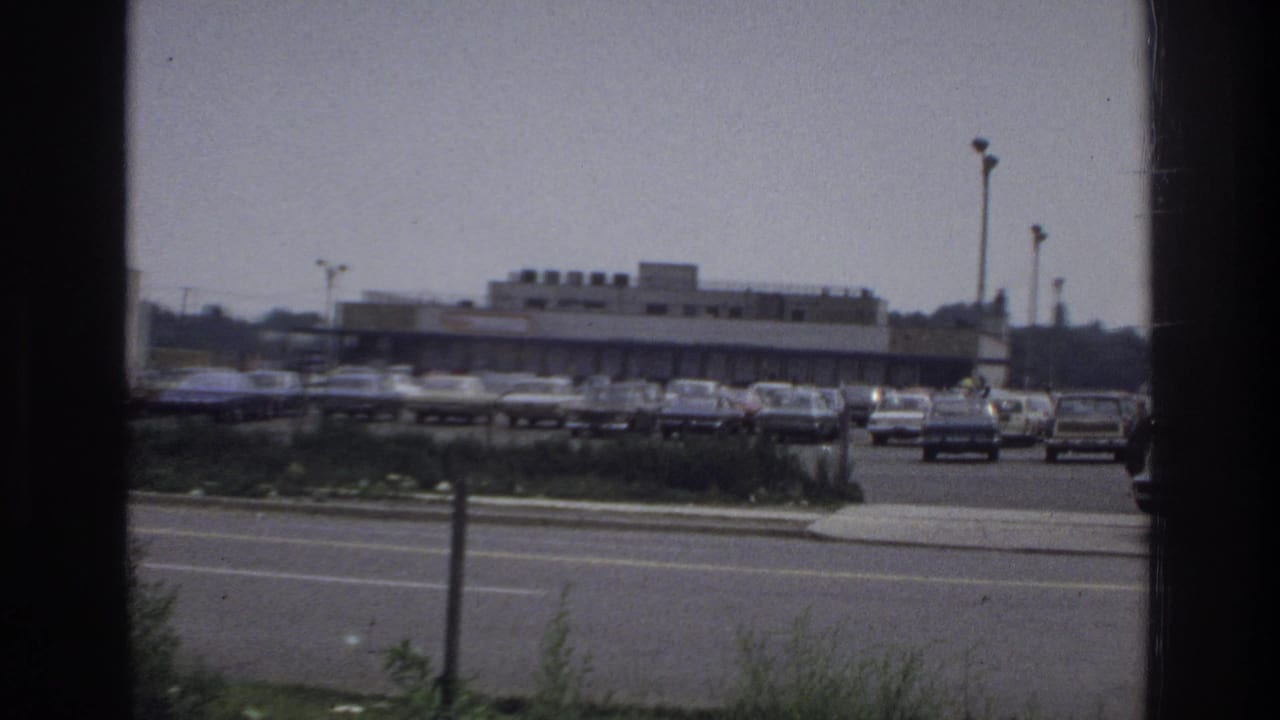 1975 Bronx shopping-center parking lot viewed through a window frame, with rows of parked cars, street traffic, light poles, and low retail buildings.