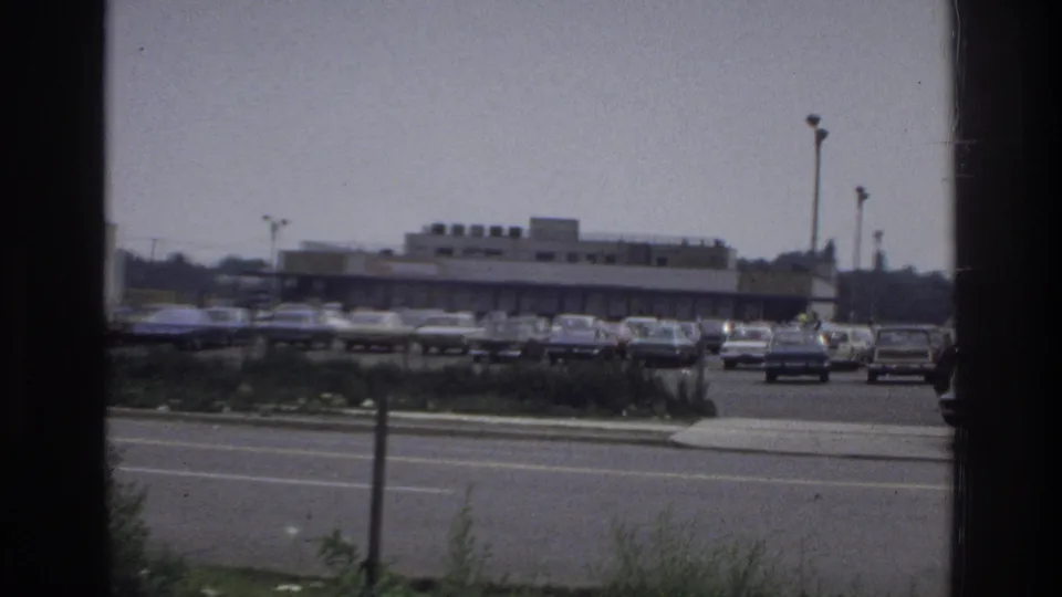 1975 Bronx shopping-center parking lot viewed through a window frame, with rows of parked cars, street traffic, light poles, and low retail buildings.