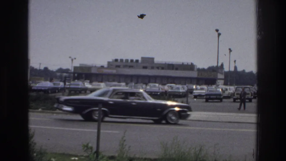 A moving 1970s sedan crosses the foreground near a Bronx shopping-center lot, with parked cars, low buildings, light poles, and a pedestrian visible.