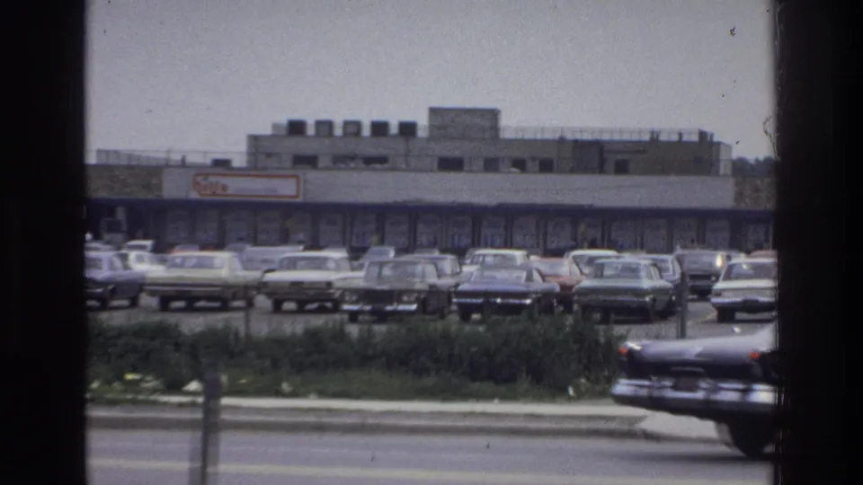 Closer 1975 Bronx view of a shopping-center facade and crowded parking lot, with period cars lined up behind roadside grass and a passing car edge.