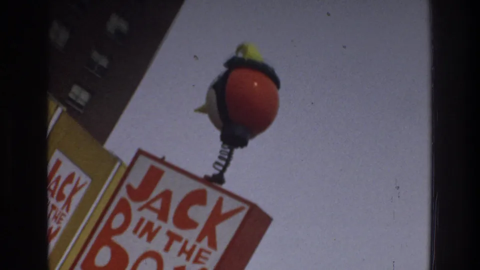 Cropped 1975 Bronx roadside view of a red-and-yellow Jack in the Box sign against the sky, with part of the sign structure and building edge visible.