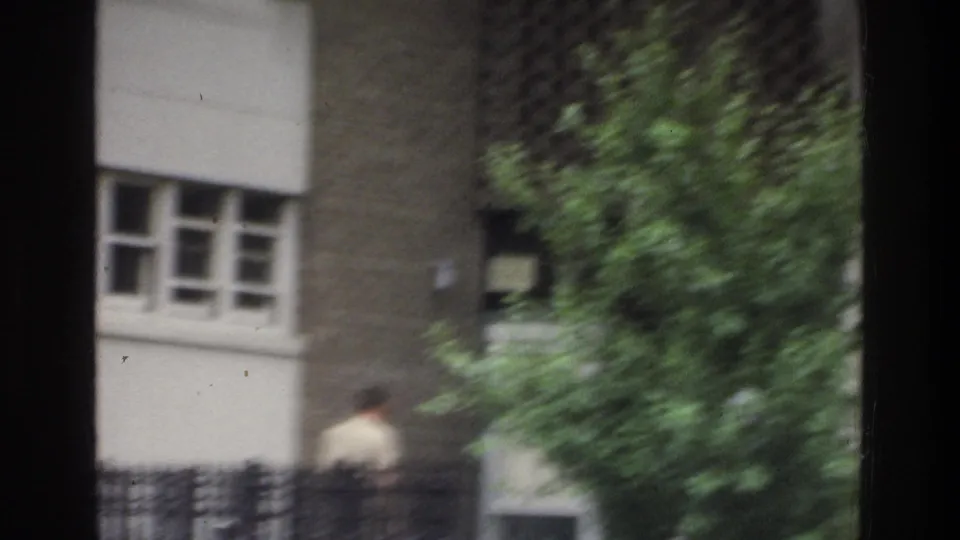 1975 Bronx 8mm frame shifting away from the sign to a soft, handheld view of a building exterior with windows, a fence, and a leafy tree.