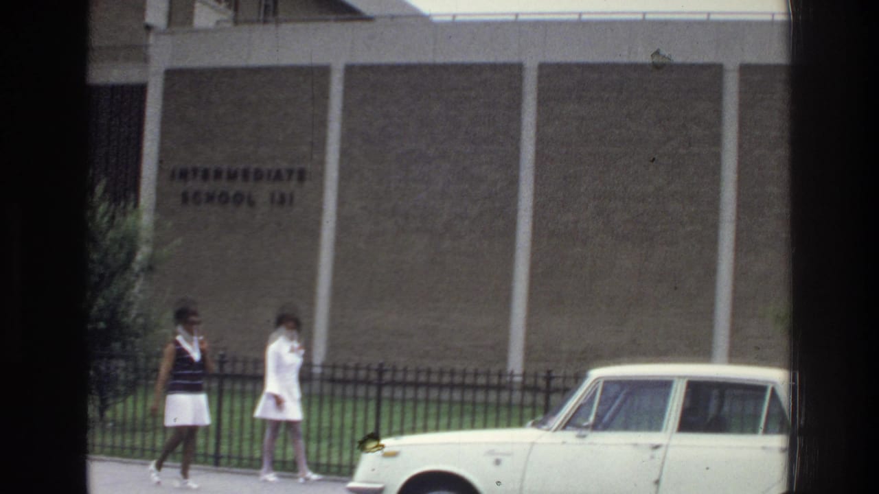 Two women walk on a sidewalk beside a school wall and a parked light-colored sedan.