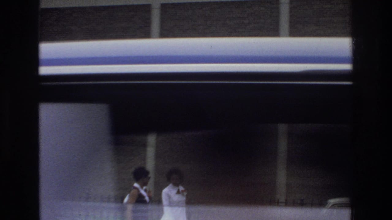 Viewed through window framing, two women appear near the Bronx school exterior with brick and concrete walls behind them.