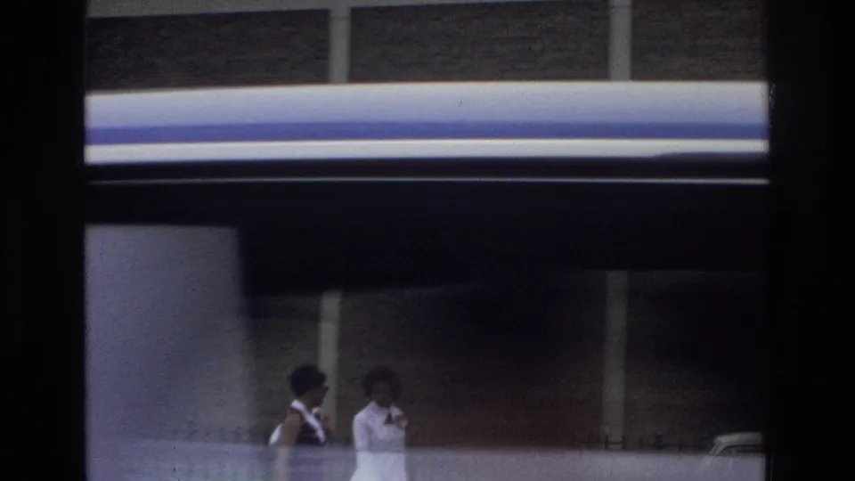 Viewed through window framing, two women appear near the Bronx school exterior with brick and concrete walls behind them.