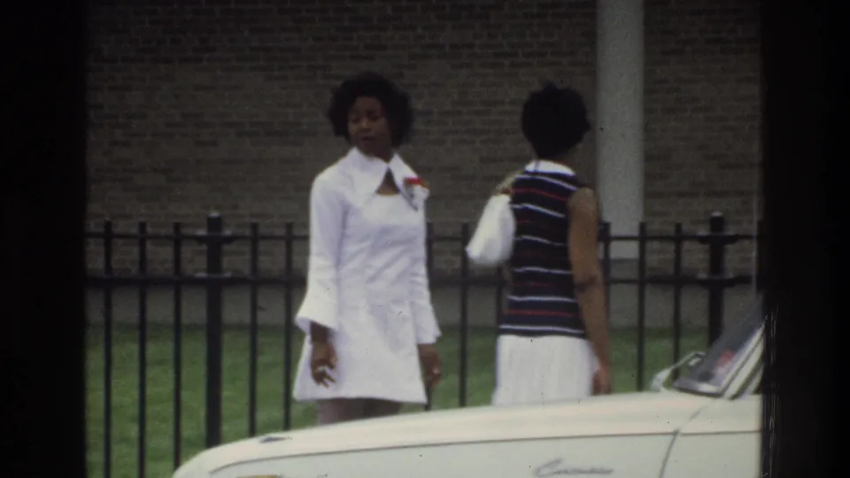 Two women near Intermediate School 131 in the Bronx are framed by a black metal fence and the hood of a light-colored sedan.