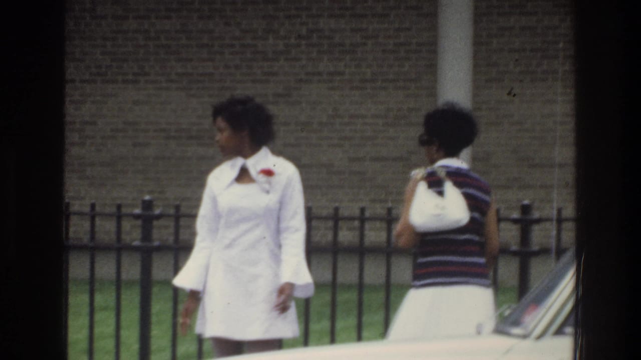 In 1975 Bronx home-movie footage, two Black women walk outdoors near a black metal fence, brick wall, and partially visible car.