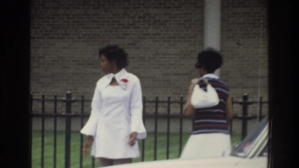 In 1975 Bronx home-movie footage, two Black women walk outdoors near a black metal fence, brick wall, and partially visible car.
