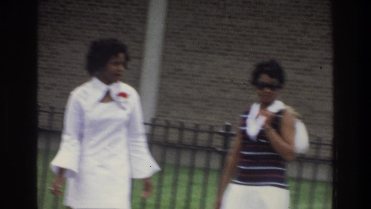Two women stand or walk beside a fence, one in a white dress and one wearing sunglasses with a white purse.