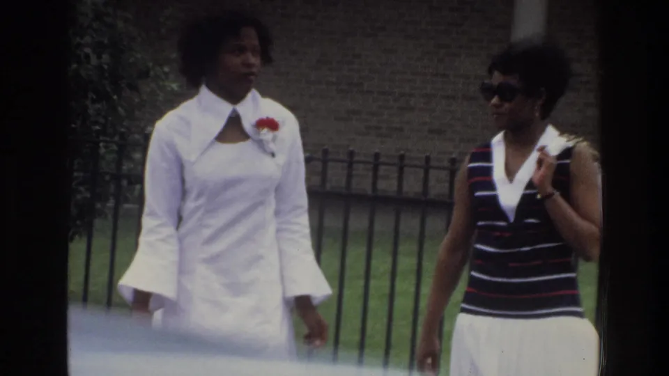 Two Black women walk outdoors in 1975 Bronx footage, framed by a metal fence, brick background, and soft foreground car blur.
