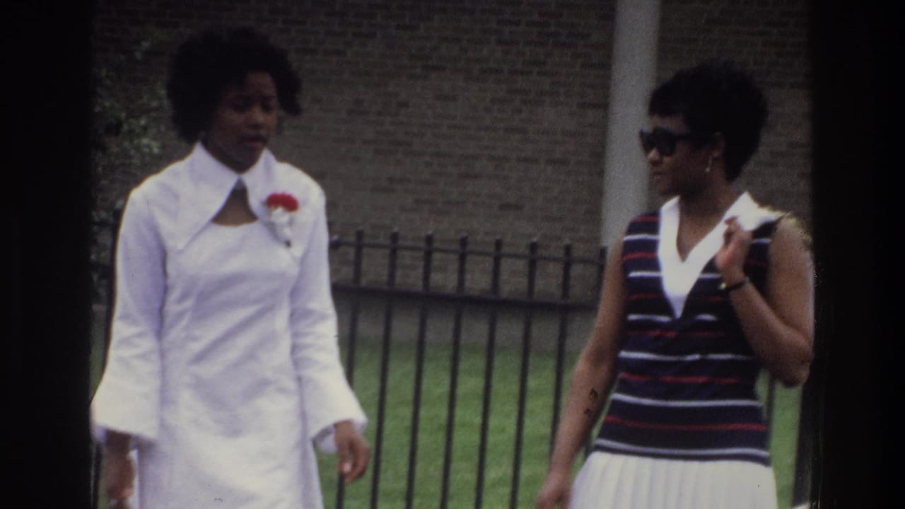 Two young Black women walking past a black iron fence and brick wall, identified as the Bronx, New York, circa 1975; one wears a long white dress with wide collar and red corsage.