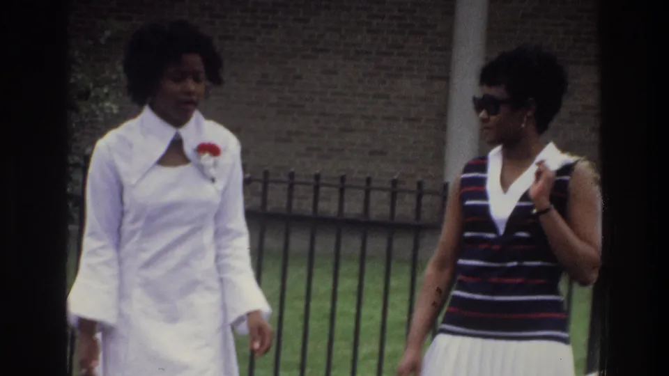 Two young Black women walking past a black iron fence and brick wall, identified as the Bronx, New York, circa 1975; one wears a long white dress with wide collar and red corsage.