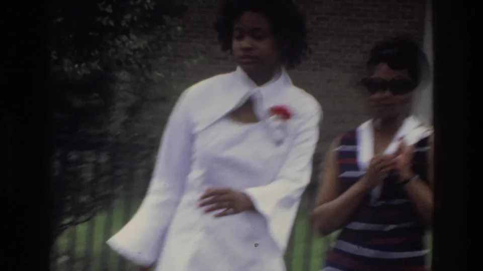 Medium-close 8mm frame of two women outdoors, identified as the Bronx, circa 1975; wide-lapel white dress and red corsage in foreground, second figure with large sunglasses behind.