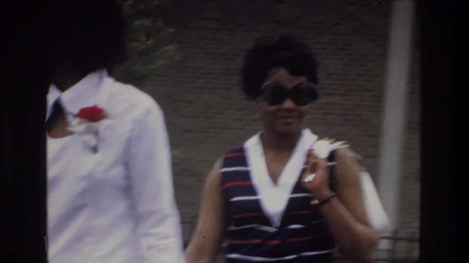 Closest 8mm frame of two women, identified as the Bronx, New York, circa 1975; woman in striped sleeveless top and round sunglasses smiles; red corsage clearly visible on white dress.