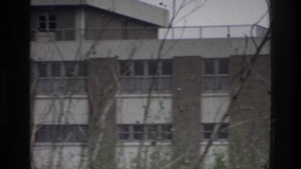 1975 Bronx home-movie frame showing a brick-and-concrete building with window rows, seen through bare branches from a moving vehicle.