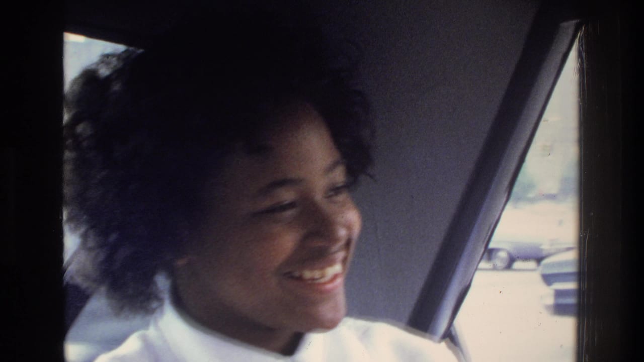 Young woman with curly hair laughing in a vehicle interior; 1970s parked cars and a building are visible through the rear window.