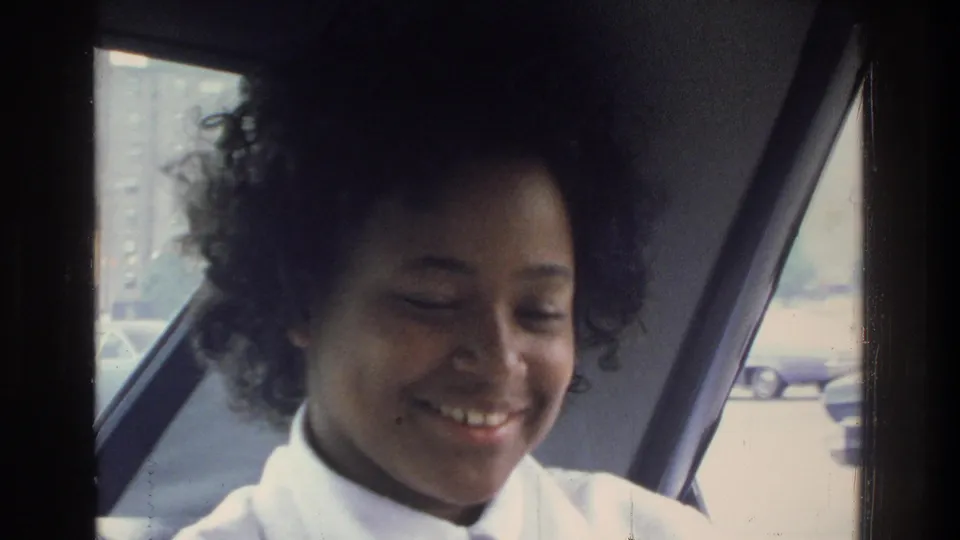 Young woman with natural curly hair smiling warmly in a vehicle; 1970s street scene with parked cars and buildings through the window; Bronx, New York, circa 1975.