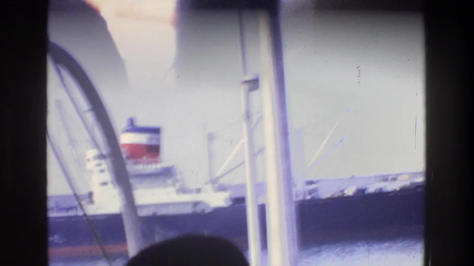 Large ocean-going vessel with red and black funnel at a New York-area harbor dock, viewed through a window with metal railings in the foreground, circa 1975.