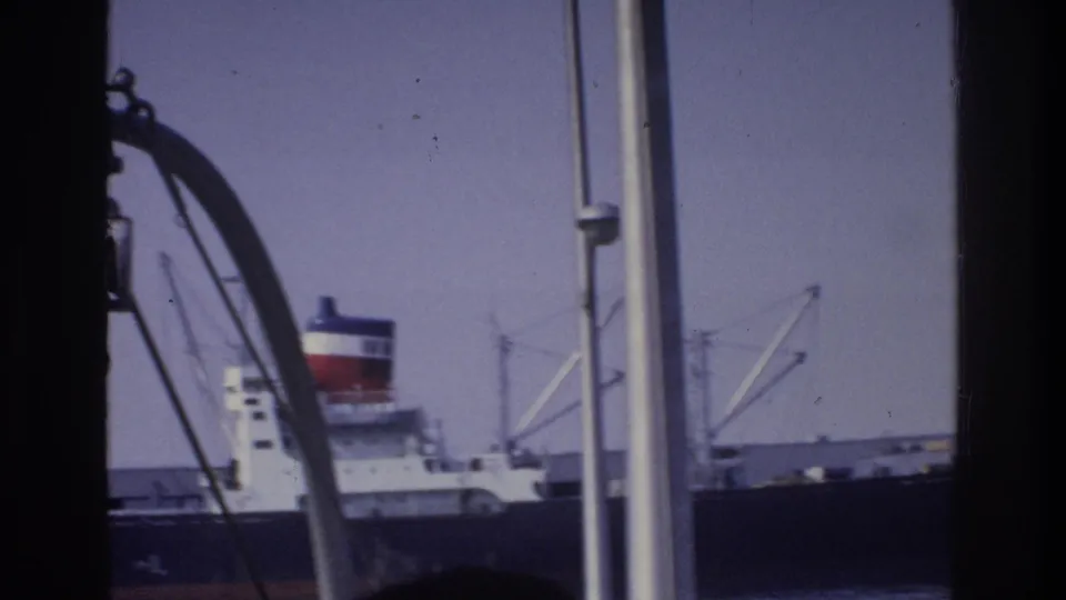 Ocean-going vessel with distinctive red and black funnel at a New York-area harbor dock, framed by metal structural elements in the foreground, circa 1975.