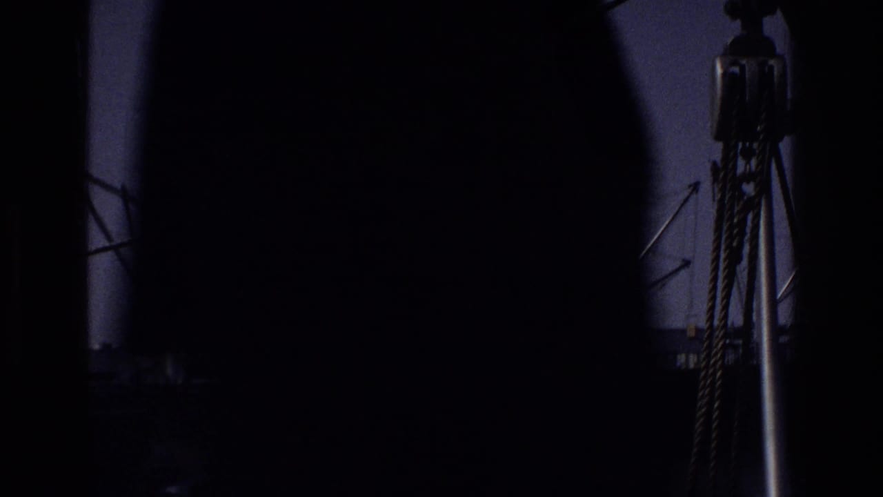 Heavily shadowed opening frame from a 1975 New York dock, showing rope or cable rigging and a partial crane arm through a near-opaque foreground silhouette.