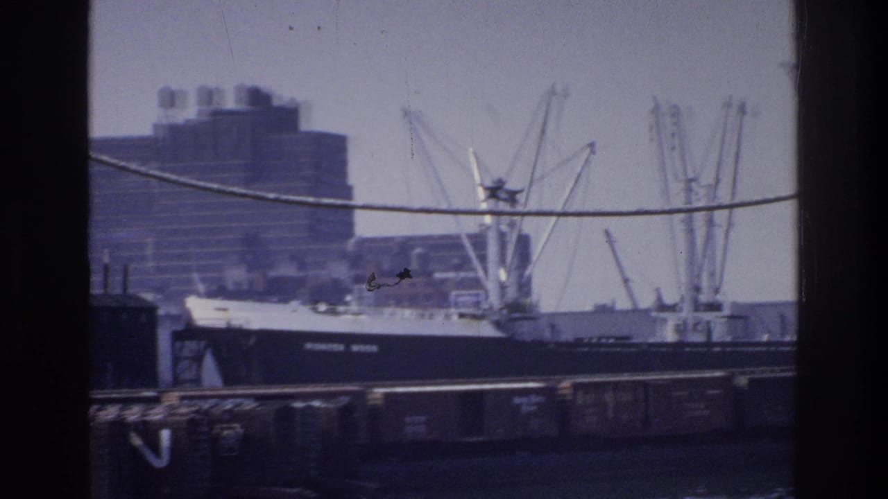 Cargo ship at dock flanked by multiple lattice cranes, a large multi-story building visible behind under overcast sky.