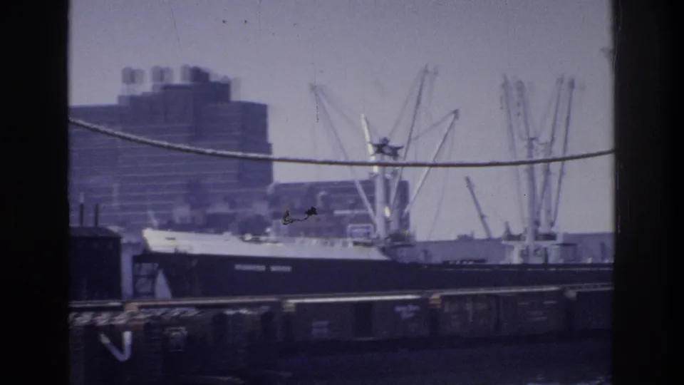 Mid-1970s cargo freighter berthed at a New York City waterfront dock, lattice-steel cranes on both sides, large urban building in background under a hazy overcast sky.