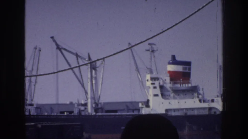 Cargo freighter with a tricolor banded funnel docked at the Bronx waterfront circa 1975, lattice cranes visible, mooring rope arcing across frame, and a dark head silhouette in the foreground.