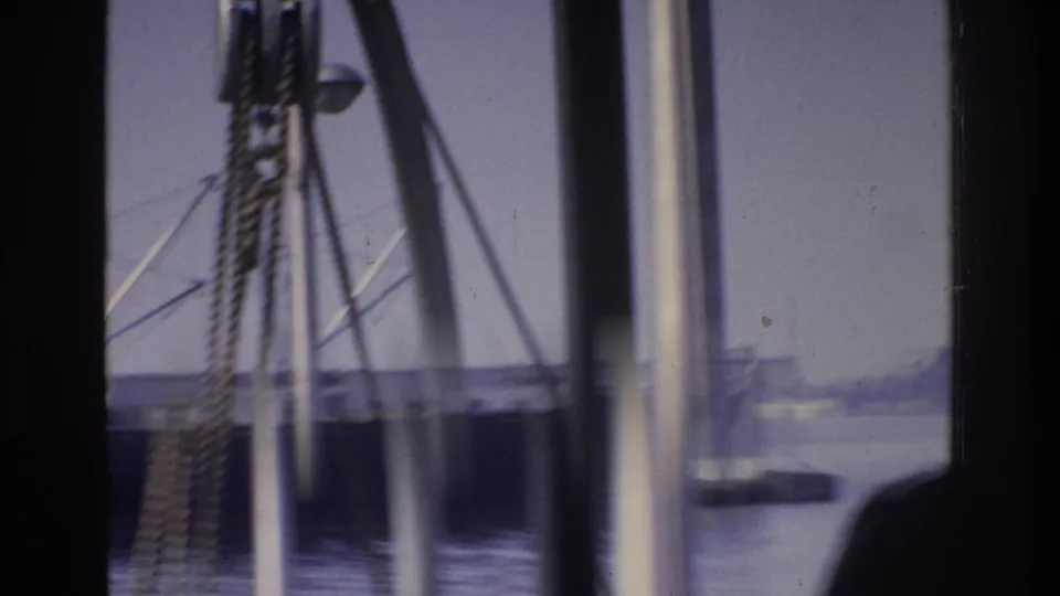 View through heavy dock mooring lines at a 1975 New York waterfront, calm water and a faint distant vessel on the horizon, with a person silhouette visible at frame edge.