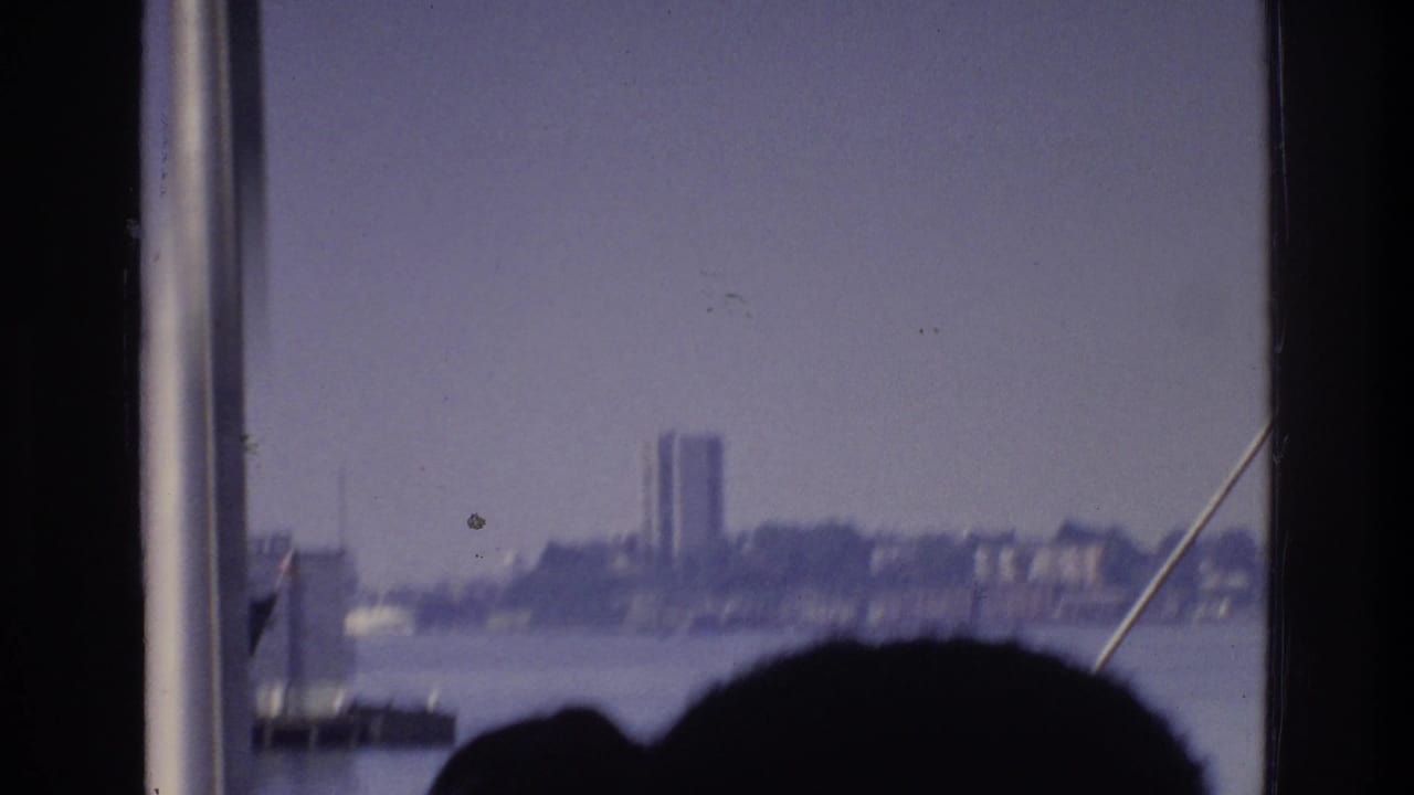 1975 8mm home-movie shot from a New York waterway boat; rigging cable and a silhouetted head frame a hazy Bronx-area industrial skyline with one prominent tall building.