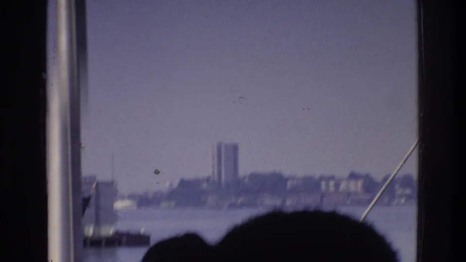 1975 8mm home-movie shot from a New York waterway boat; rigging cable and a silhouetted head frame a hazy Bronx-area industrial skyline with one prominent tall building.
