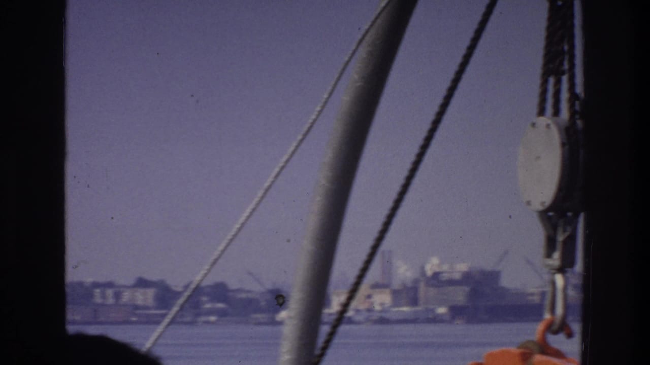 Boat rigging cables and a metal pulley fitting cross the frame; industrial buildings line the far shore across calm water; orange safety equipment visible at right edge.