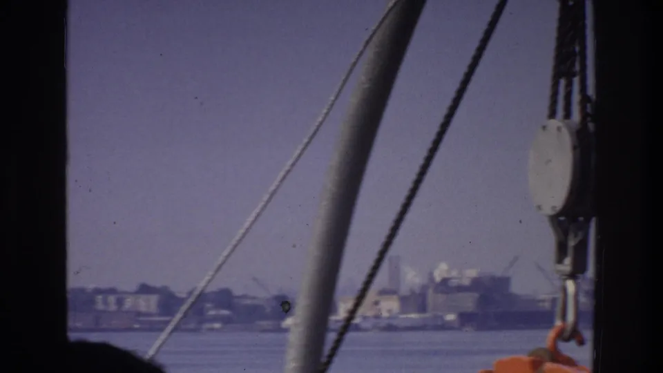 8mm frame from a 1975 New York boat trip; diagonal rigging cables, a metal pulley fitting, and orange life-ring edge frame a hazy industrial waterfront across the water.