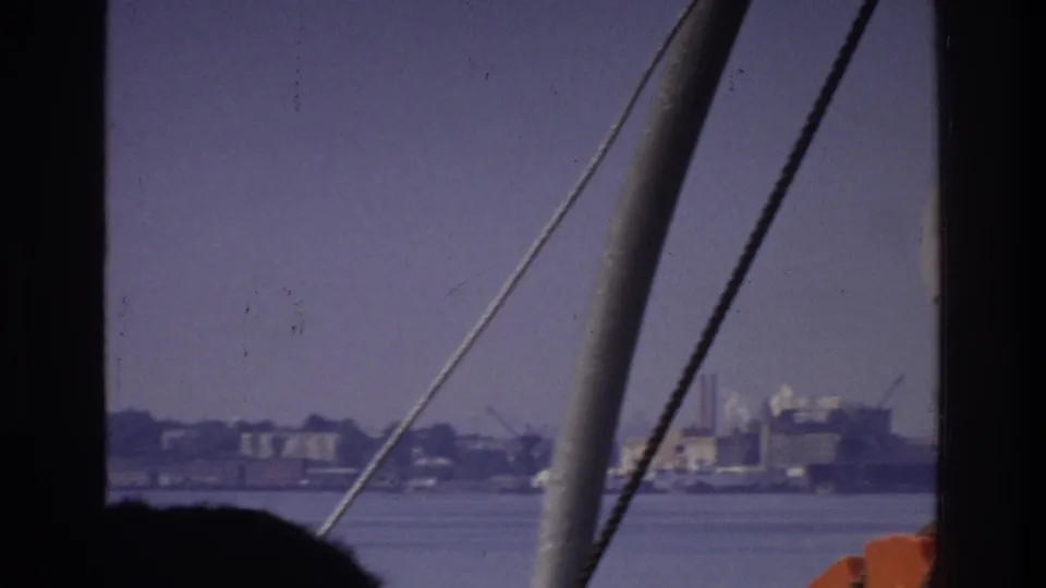 8mm home-movie frame circa 1975; boat rigging cables and an orange life-ring frame a hazy industrial waterfront with dock structures across the water channel.