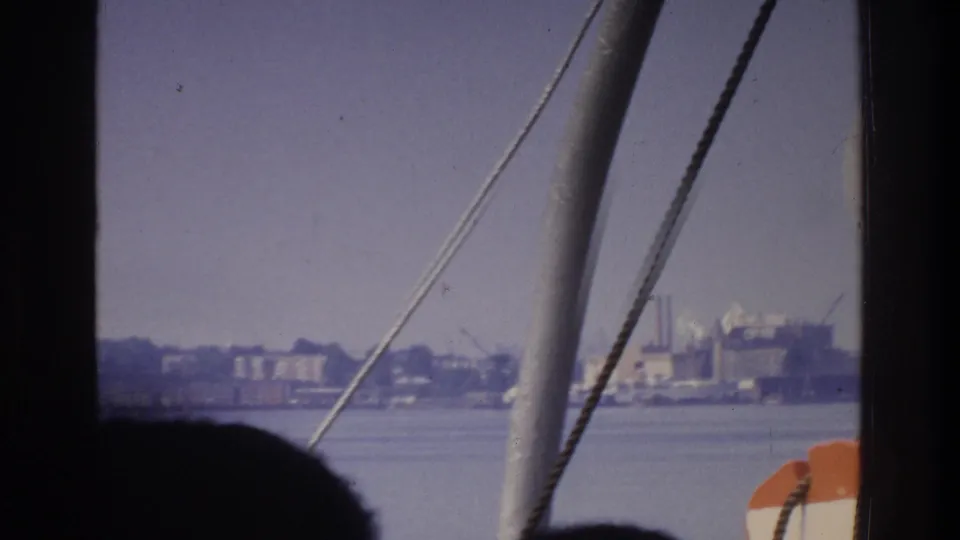 Final 8mm frame from a 1975 New York waterway boat trip; rigging cables, orange life-ring, and a silhouetted figure frame a hazy low-rise industrial waterfront.