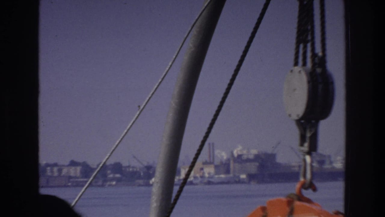 Boat rigging with a pulley block and hook visible at right, orange equipment at frame edge, and an industrial waterfront across water.