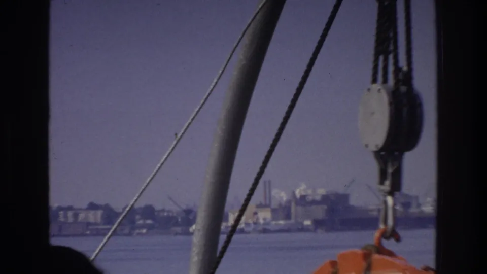 8mm footage circa 1975 from a vessel showing nautical rigging hardware including a pulley block and orange safety equipment, with an industrial shoreline in the background.