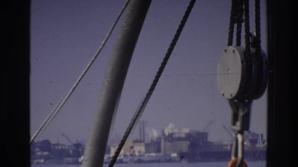 8mm home movie circa 1975 with a prominent block-and-tackle pulley in the mid-foreground and a low industrial shoreline across a harbor or river behind it.