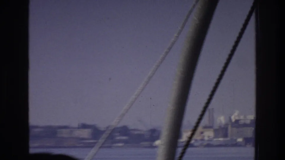8mm home movie circa 1975 showing boat rigging in the foreground and a distant industrial shoreline — possibly New York — with characteristic purple emulsion color shift.