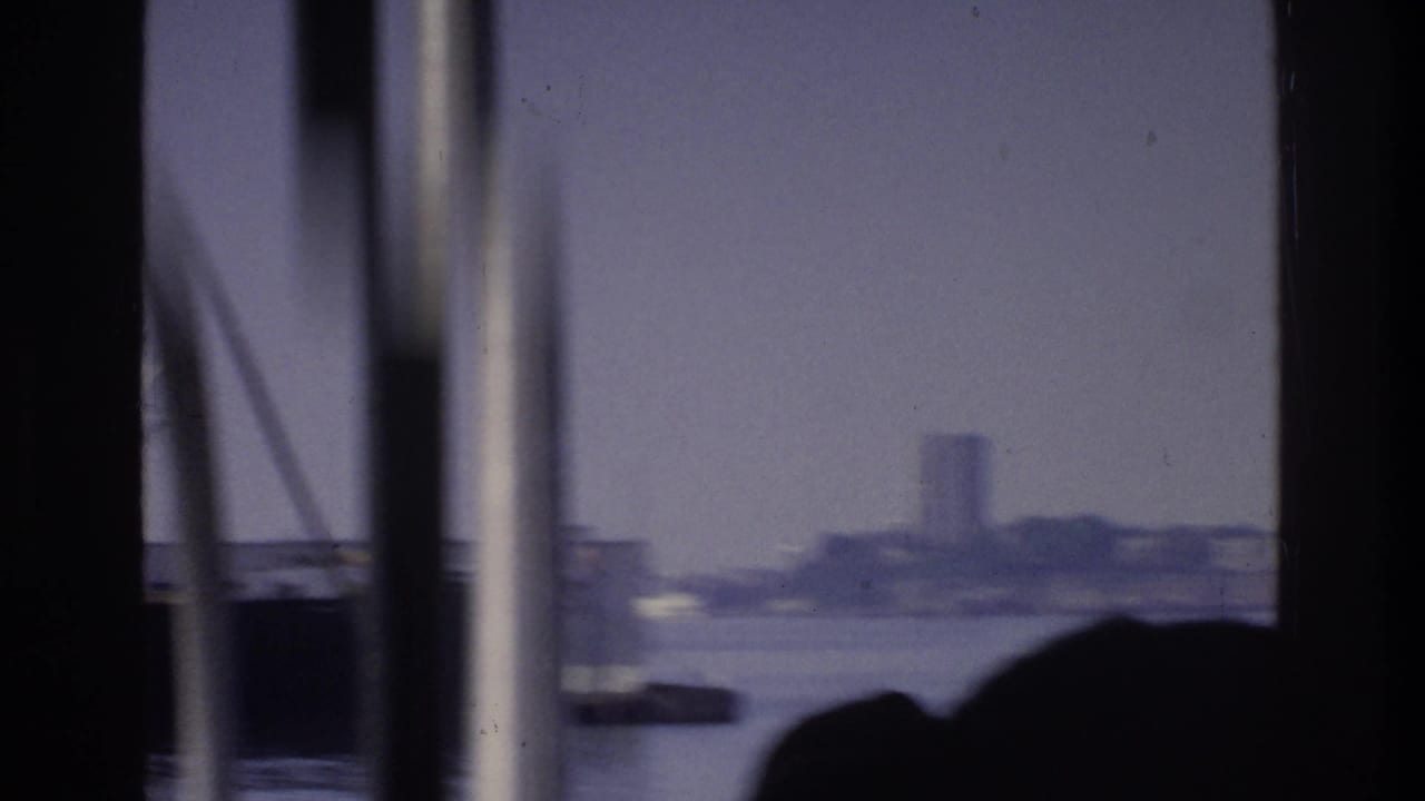 8mm opening frame from a boat in New York harbor, circa 1975; rigging poles and two silhouetted passengers frame a distant ship on the water.