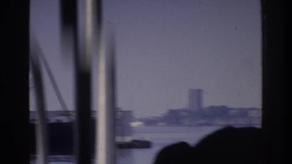 8mm opening frame from a boat in New York harbor, circa 1975; rigging poles and two silhouetted passengers frame a distant ship on the water.