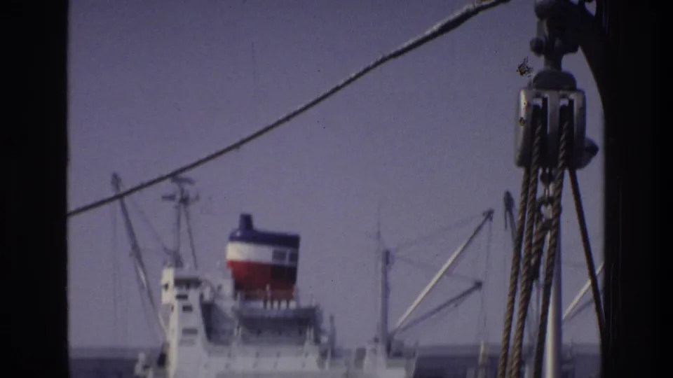 Large white cargo ship with tricolor funnel viewed through harbor rigging from a passing boat, New York harbor area, circa 1975; faint urban skyline behind.