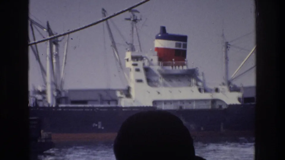 Close harbor view of white cargo ship with tricolor funnel from a moving boat, circa 1975 New York; foreground rigging cable and silhouetted observer at bottom.