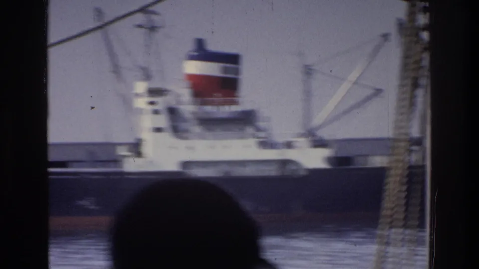 8mm shot of white cargo ship with tricolor funnel in New York harbor, circa 1975; rope rigging at right edge, observer's silhouette at lower frame.