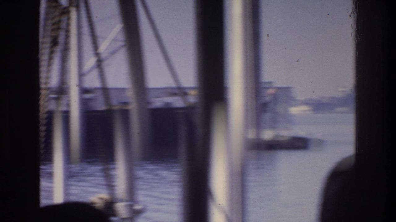 8mm shot circa 1975, looking across a New York waterway from a moving boat; rigging poles visible in foreground, low buildings on far shore.