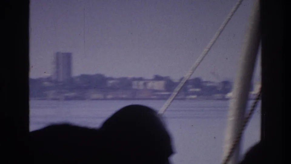 8mm home movie circa 1975; silhouetted boat passenger in foreground, modernist high-rise tower visible across a New York waterway.