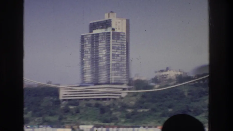 8mm footage circa 1975; multi-story modernist residential tower on a vegetated hillside, viewed from a moving boat on a New York waterway.