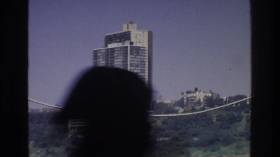 8mm home movie circa 1975; a boat passenger's silhouette dominates the foreground while a modernist tower and hillside buildings appear across the New York waterway.