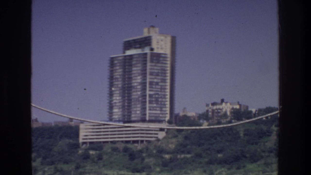 8mm film frame circa 1975, Bronx, New York — glass high-rise tower and lower stepped building seen from moving vehicle with cable in foreground.